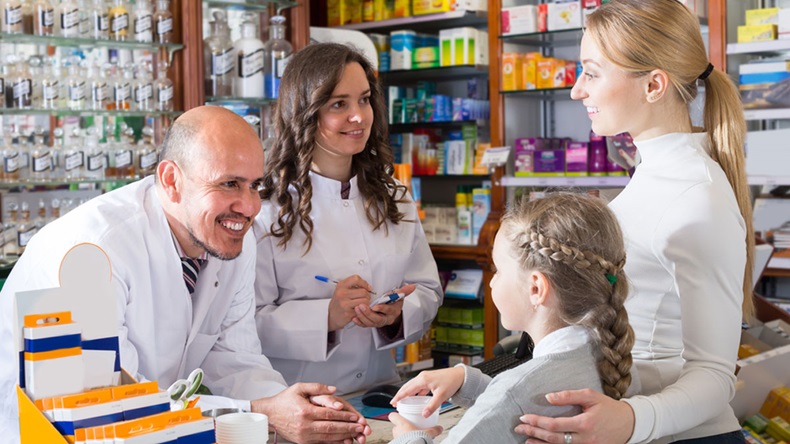 Two friendly smiling pharmacists wearing white coats standing next to cashier and consulting a customer with child. - Image 