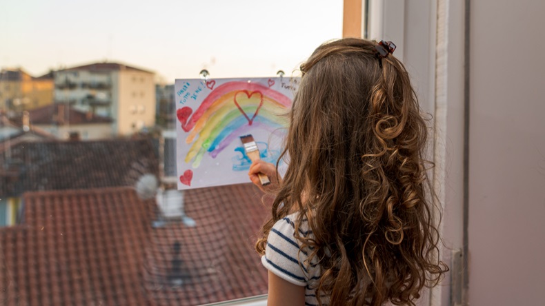 little girl is painting a rainbow to a sheet of paper attached on her window during the covid-19 quarantine in italy