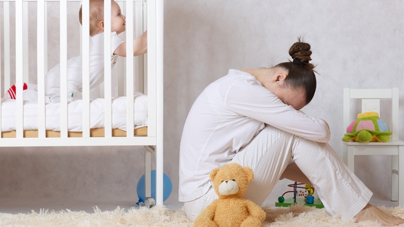 woman sitting on floor with head down next to baby in crib