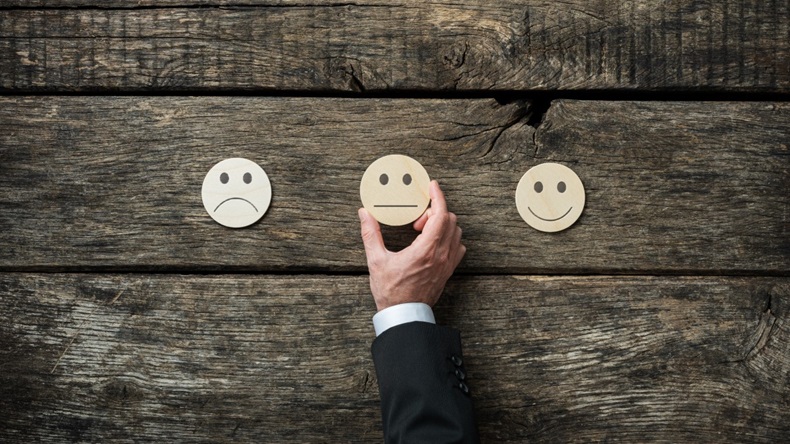 Wooden background with three drawn emoticon images in middle: sad, neutral and happy. Hand reaching out to place neutral emoticon. 